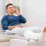 Man with a leg in plaster sitting on a sofa at home with crutches nearby and making a phone call