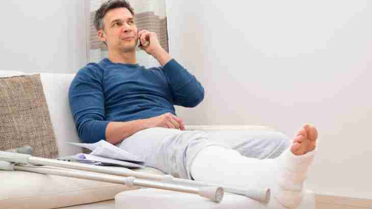 Man with a leg in plaster sitting on a sofa at home with crutches nearby and making a phone call