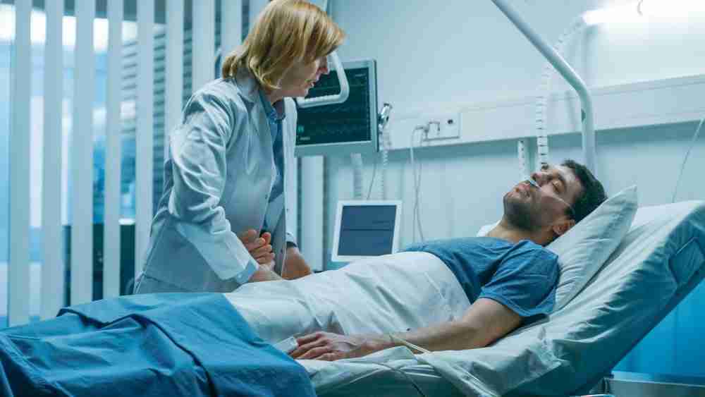 Death Compensation Claim Man lying unconscious in a hospital bed connected to life support with a woman holding his hand