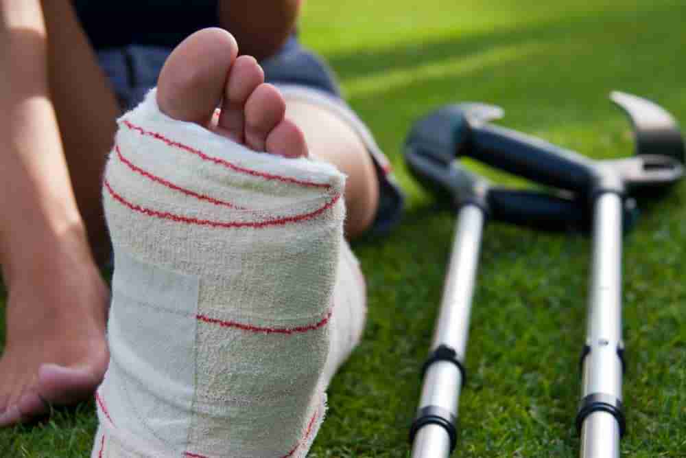 Person with a foot in plaster sitting on grass with a crutch beside the injured leg