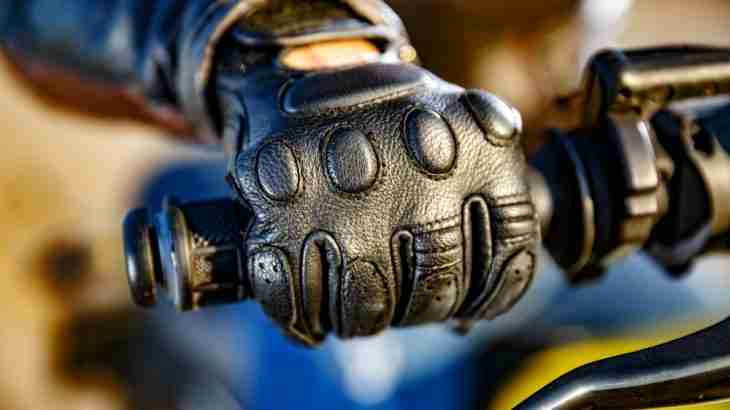 Close-up of a gloved hand on a motorcycle handlebar with a rider and motorcycle partly visible in the background